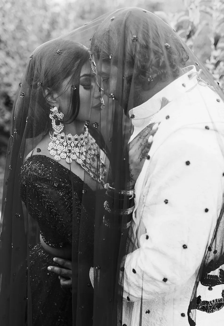 Black-and-white portrait of the couple embracing beneath a beaded veil